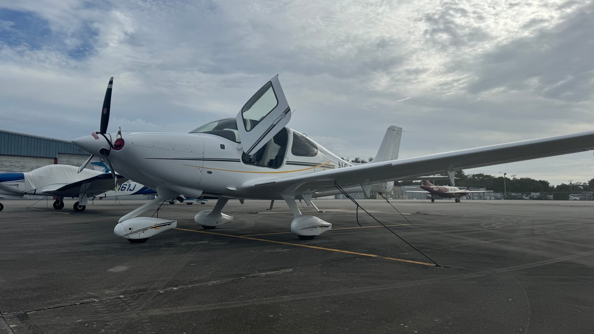 A white Cirrus SR20 with a gold pinstripe parked on the ramp with its cabin door open, other aircraft and a hangar behind it under an overcast sky